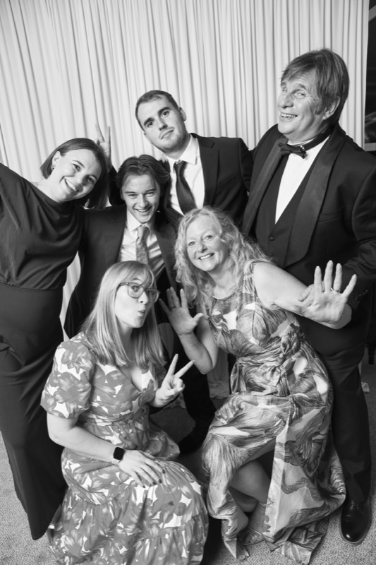 A family of six in evening wear pose together, waving and pulling faces